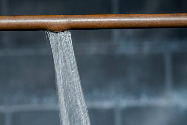 A close-up of a copper pipe that ruptured because it had frozen, now spraying water, with a cinder block wall in the background.